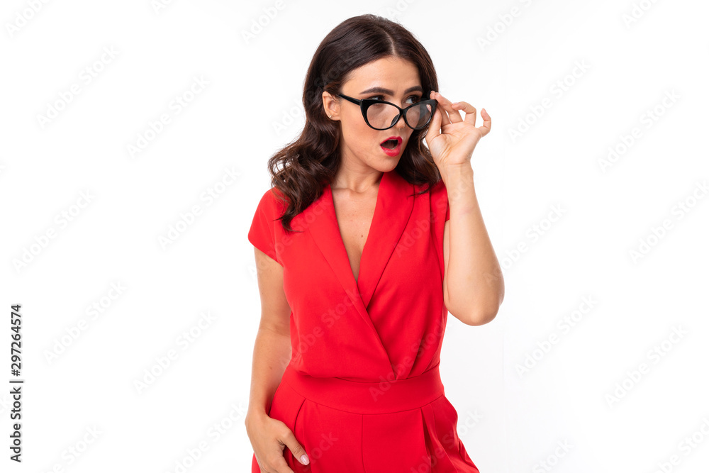 A young woman with red lips, bright makeup, dark wavy long hair, in a red suit, black glasses with transparent stacks stands and is shocked