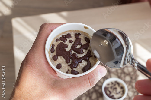A men eats healthy raw ice cream with spoon. Top view from myself. delicious fruit vegan dessert in a white paper cup in a male hand. A small spoon in the other hand. Kitchen table in background