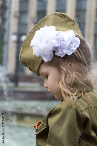 Little girl dressed in military uniform on background of fountains near the government building. White bow on her head. Children of war. Defenders of the Fatherland. Victory Day celebrations. Profile