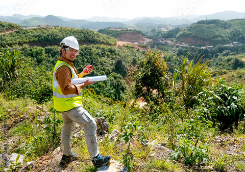 Engineer manager man with white helmet and hold construction paper plan and fm handheld transceiver or walkie talkie look back to camera and stand in front of mountain.