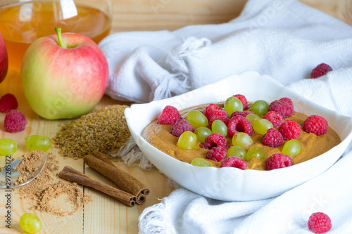 Healthy meal concept background. Vegan flax porridge with raspberry and grape raisins in white bowl on fabric. Apple, honey, cinnamon sticks and powder on wooden table. Delicious raw foods breakfast