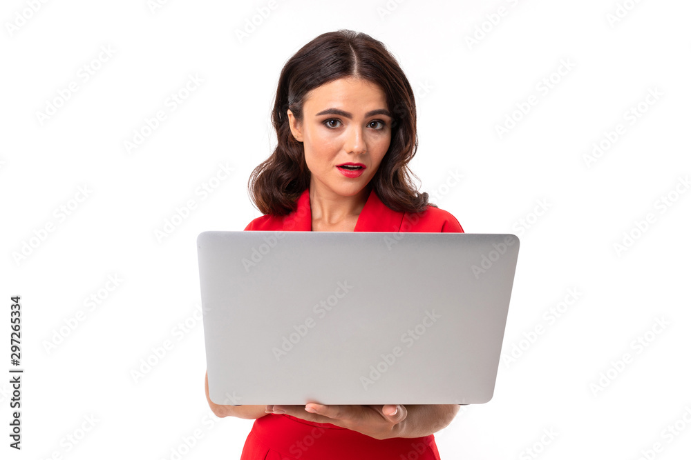 A young woman with red lips, bright makeup, dark wavy long hair, in a red suit, stands with a white laptop A young woman with red lips, bright makeup, dark wavy long hair, in a red suit, stands with a white laptop