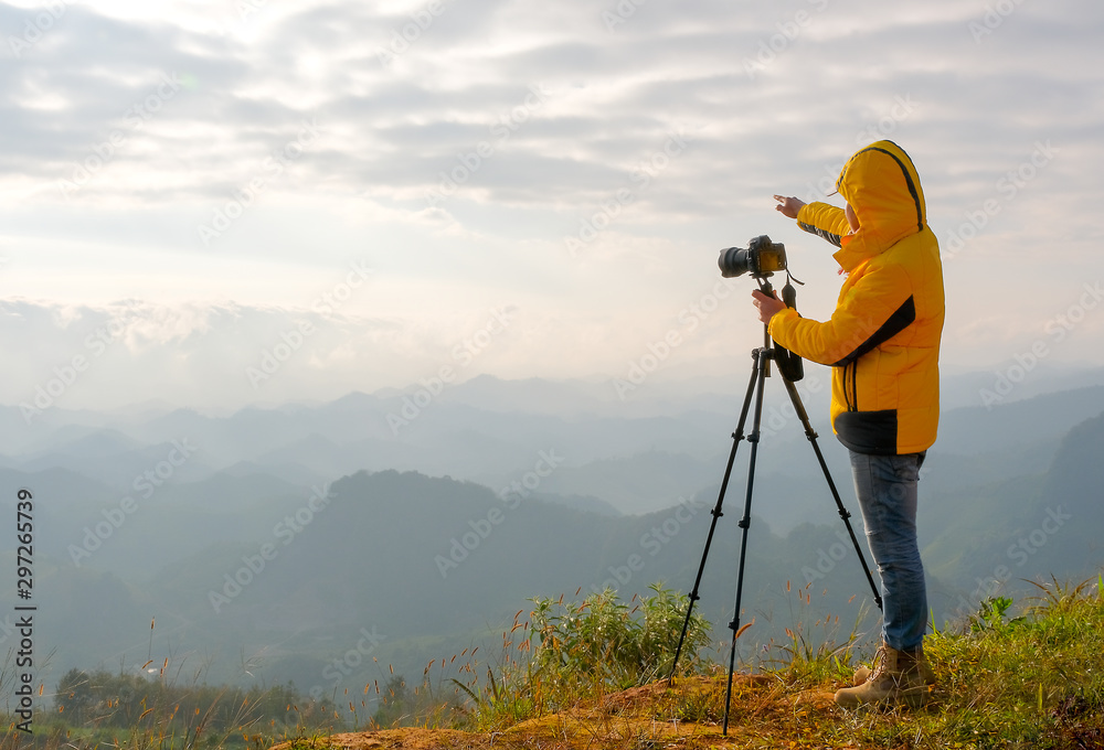 The photographer with yellow jacket stand near to his camera and stand in front cliff, cloudy and point to in front direction.
