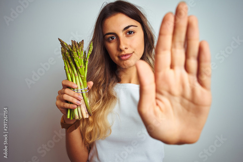 Young beautiful woman eating asparagus over grey isolated background with open hand doing stop sign with serious and confident expression  defense gesture