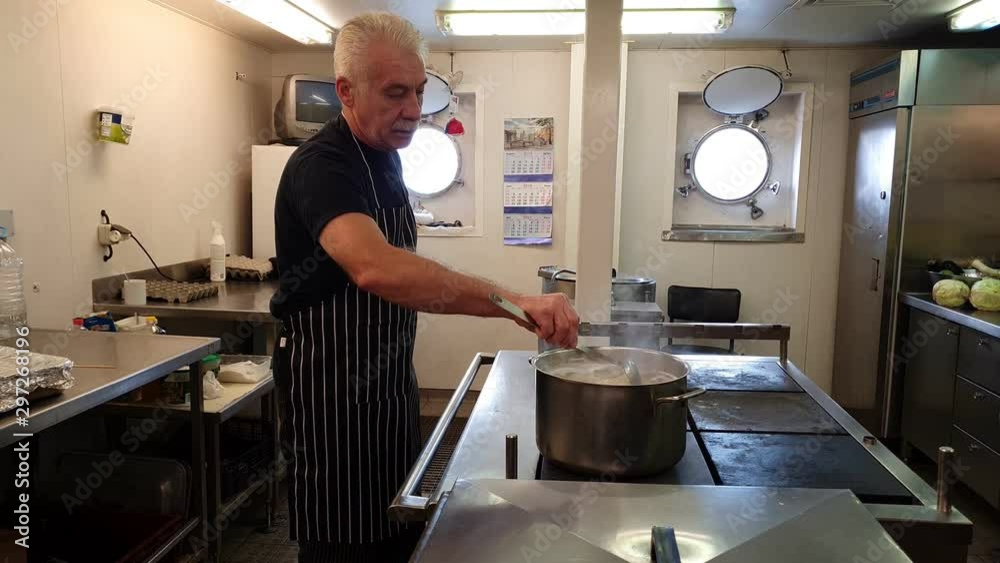 professional senior cook in black striped apron mixes boiling dish in metal saucepan in oil tanker kitchen professional senior cook in black striped apron mixes boiling dish in metal saucepan in oil tanker kitchen