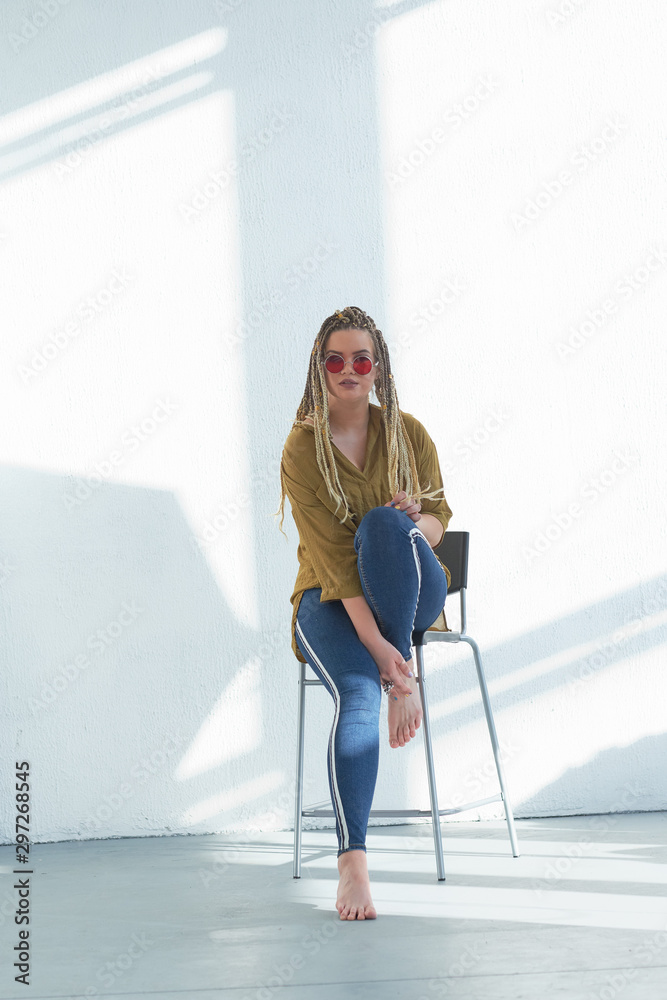 Cute lovely young woman in round sunglasses sits on a chair in the studio. against a white wall Cute lovely young woman in round sunglasses sits on a chair in the studio. against a white wall
