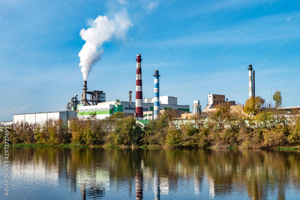 Obraz premium pipes of woodworking enterprise plant sawmill near river. Air pollution concept. Industrial landscape environmental pollution waste of thermal power plant