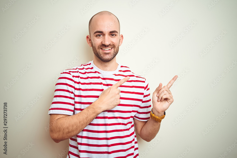 Young bald man with beard wearing casual striped red t-shirt over white isolated background smiling and looking at the camera pointing with two hands and fingers to the side. Young bald man with beard wearing casual striped red t-shirt over white isolated background smiling and looking at the camera pointing with two hands and fingers to the side.