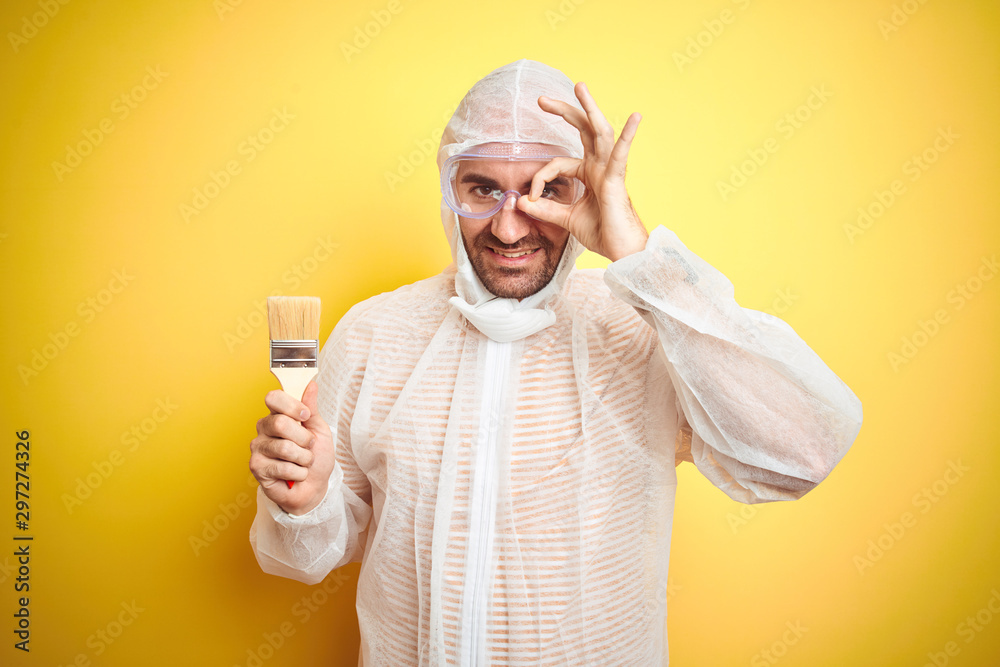 Young man wearing painter equipment and holding painting brush over isolated yellow background with happy face smiling doing ok sign with hand on eye looking through fingers Young man wearing painter equipment and holding painting brush over isolated yellow background with happy face smiling doing ok sign with hand on eye looking through fingers