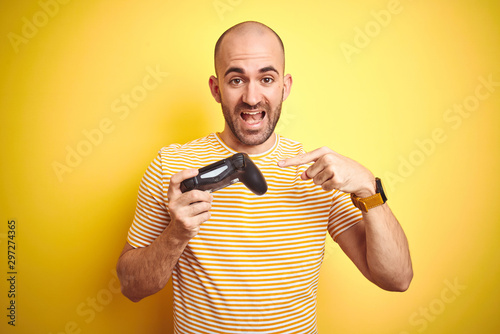 Young man playing video games using joystick gamepad over isolated yellow background very happy pointing with hand and finger