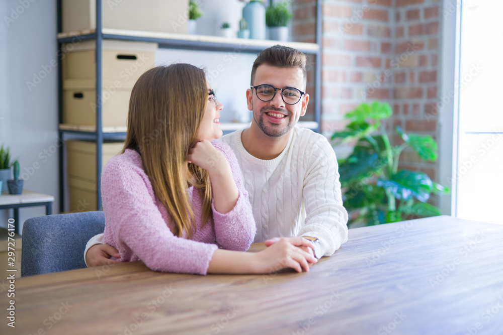 Young beautiful couple sitting on the table at home, hugging in love very happy for moving to new home with cardboard boxes behind them