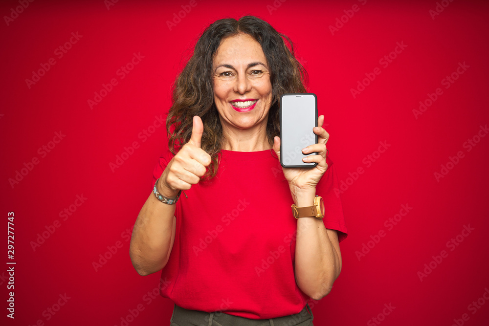 Naklejka premium Middle age senior woman showing screen of smartphone over red isolated background happy with big smile doing ok sign, thumb up with fingers, excellent sign