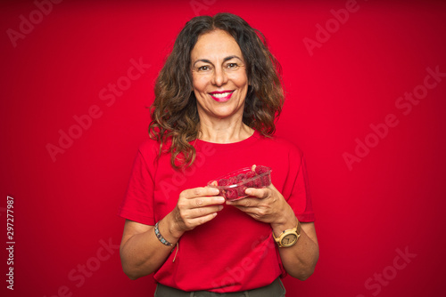Middle age senior woman eating raspberries over red isolated background with a happy face standing and smiling with a confident smile showing teeth