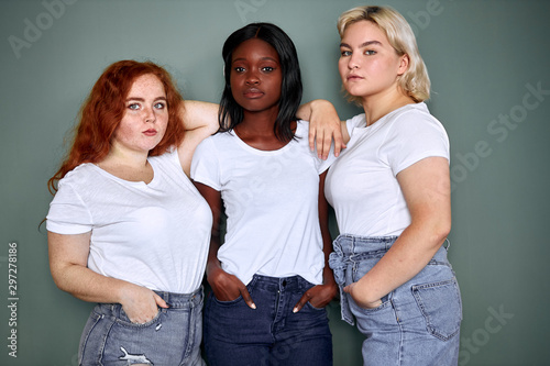 Young girls of caucasian, american and african nations. Stand together, wearing white t-shirt and jeans. Isolated over grey background. People relationship, models, fashion
