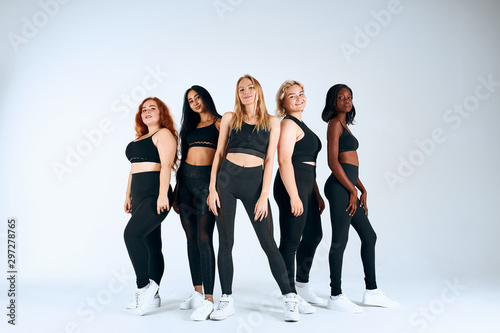 Young and positive girls of different nations stand smiling, look at camera isolated in white studio background