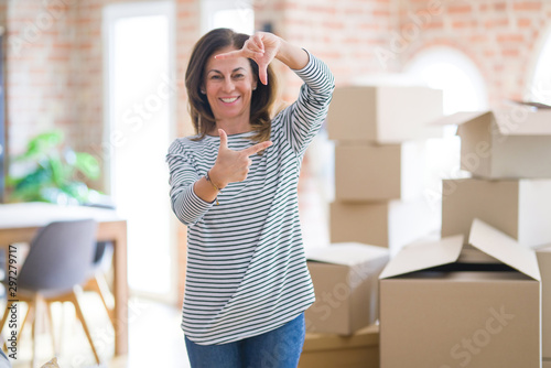 Middle age woman moving to a new house arround cardboard boxes smiling making frame with hands and fingers with happy face. Creativity and photography concept.