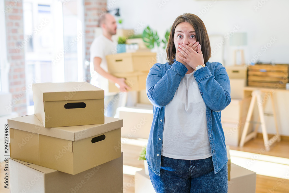 Young couple arround cardboard boxes moving to a new house, plus size woman standing at home shocked covering mouth with hands for mistake. Secret concept. Young couple arround cardboard boxes moving to a new house, plus size woman standing at home shocked covering mouth with hands for mistake. Secret concept.