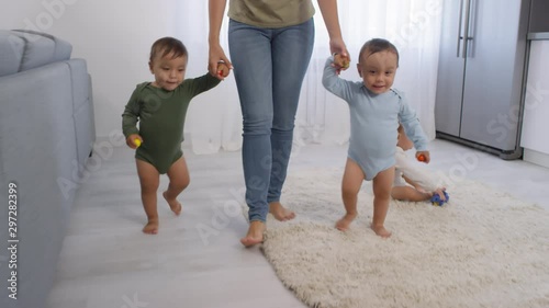 Tracking shot of unrecognizable woman and toddler twin boys in bodysuits walking together on fluffy carpet at home and holding them by hands, while their sibling is sitting and playing with toy car photo