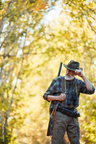 Caucasian older hunter stand in autumn forest, look away, straighten cowboy hat.