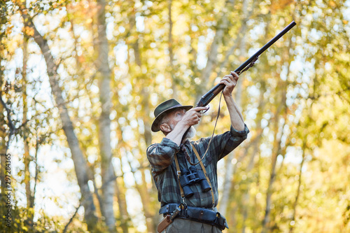 Senior hunter man aims at trophy bird in autumn forest, pointing gun at bird.