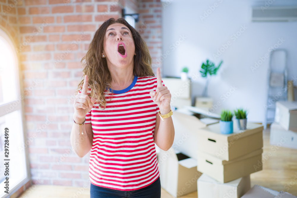 Middle age senior woman moving to a new house packing cardboard boxes amazed and surprised looking up and pointing with fingers and raised arms.