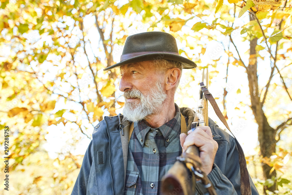 Bearded senior hunter wearing hat stands with gun on shoulder, looks into the distance in search of trophy. Wild animals prey in hunting