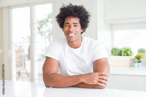 Young african american man wearing casual white t-shirt sitting at home winking looking at the camera with sexy expression, cheerful and happy face.