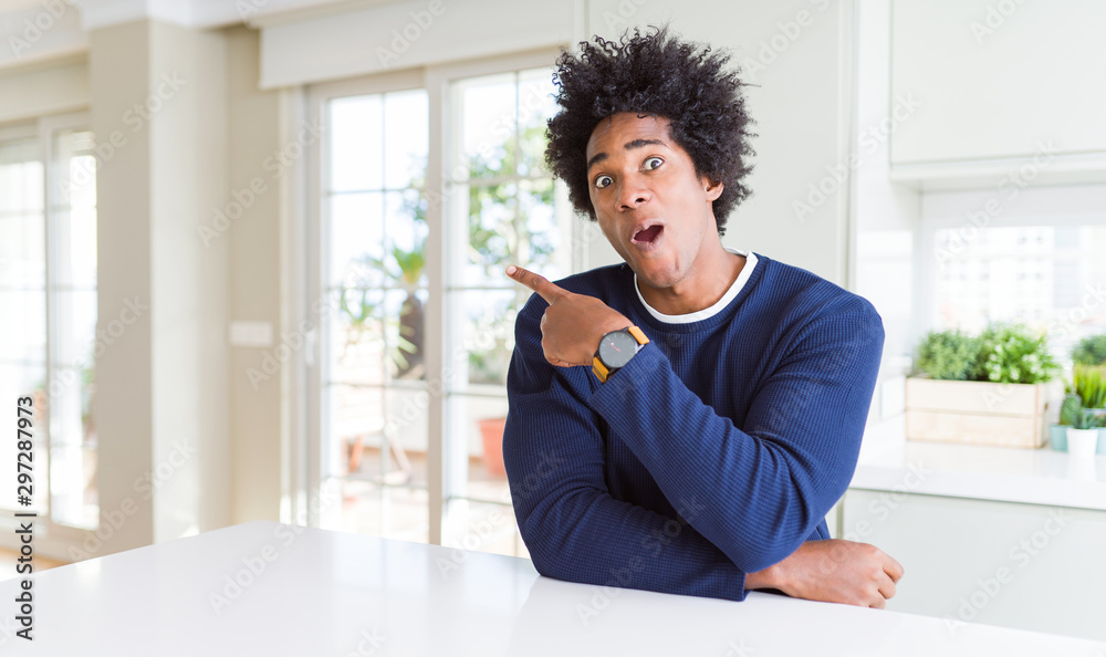 Young african american man wearing casual sweater sitting at home Surprised pointing with finger to the side, open mouth amazed expression.