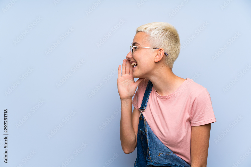 Teenager girl with overalls on blue wall shouting with mouth wide open Teenager girl with overalls on blue wall shouting with mouth wide open