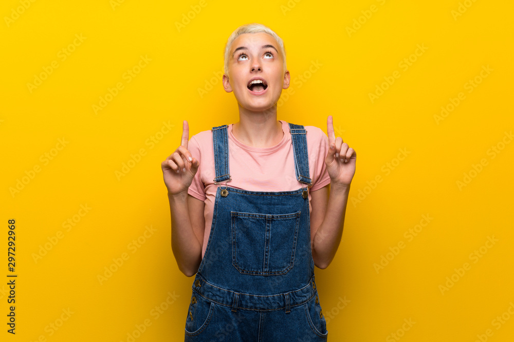 Teenager girl with overalls on yellow background surprised and pointing up Teenager girl with overalls on yellow background surprised and pointing up