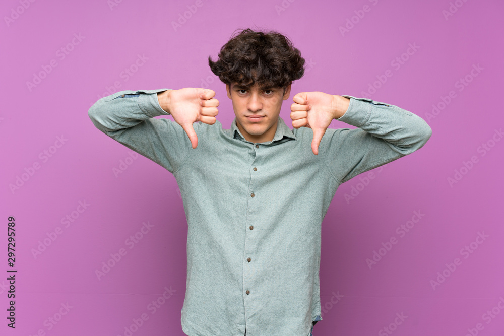 Young man over isolated purple wall showing thumb down
