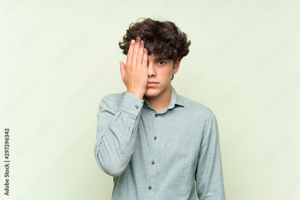 Young man over isolated green wall covering a eye by hand Young man over isolated green wall covering a eye by hand