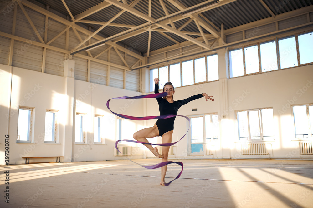 Active beautiful rhythmic gymnast standing in Sur le cou-de-pied pose, waving long colourful ribbon in the air, dancing in bright sports hall, professional sport concept - obrazy, fototapety, plakaty 