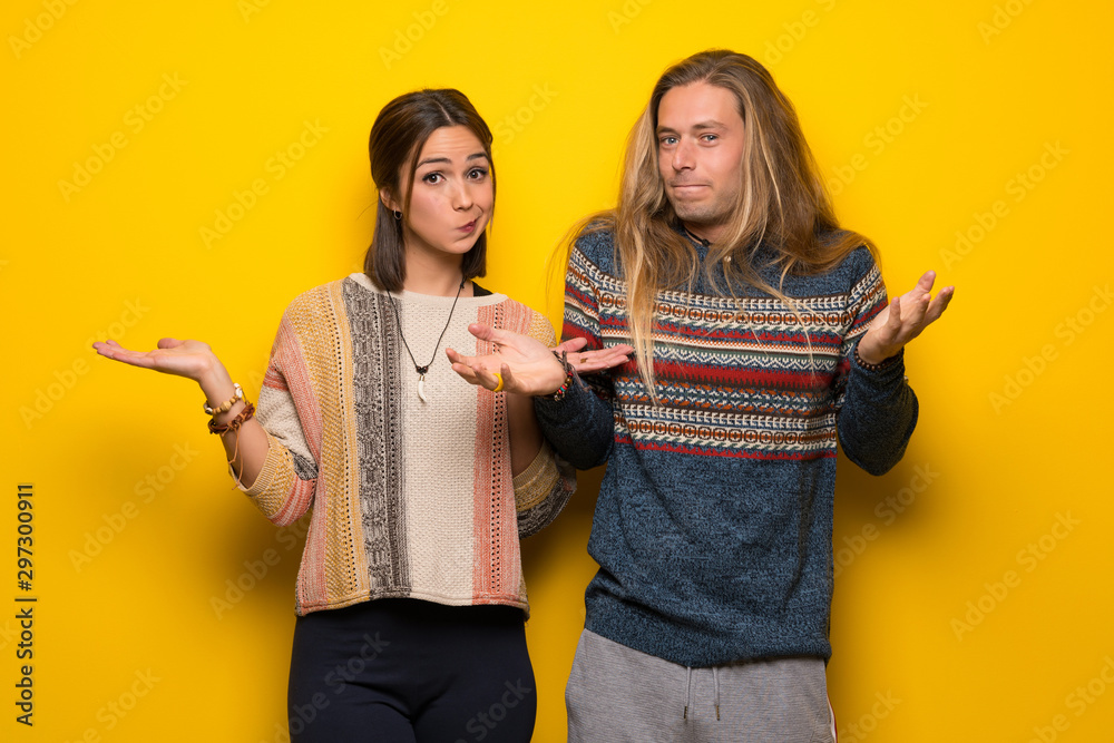 Hippie couple over yellow background having doubts while raising hands and shoulders - obrazy, fototapety, plakaty 