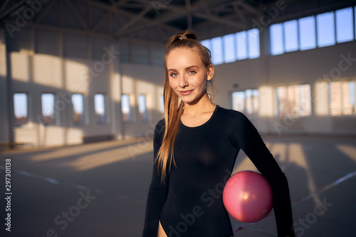 Portrait of charming beautiful woman looking straight with pleasant smile, holds pink ball, stans in dark gym hall, in gymnastics school