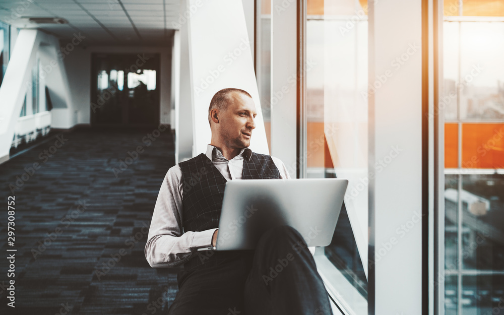 Fototapeta premium A confident serious prosperous caucasian man entrepreneur is sitting with a laptop on a white bench in a passageway of a modern business office skyscraper and pensively looking outside the window