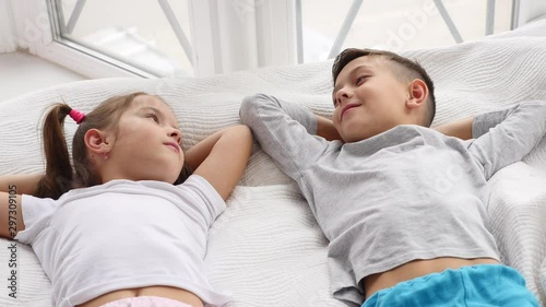 Top view on two beautiful siblings lying on a bed together, smiling and talking while having a rest.