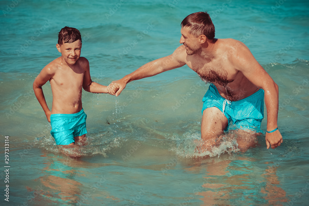 Dad and son in similar swimming shorts having fun at  beautiful seashore. They holding hands and enjoying their perfect vacations.