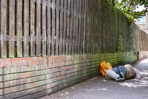 Adult male seen laying asleep on a footpath in a shaded area of an English town. He can be seen with orange shopping bags which are by a wall.