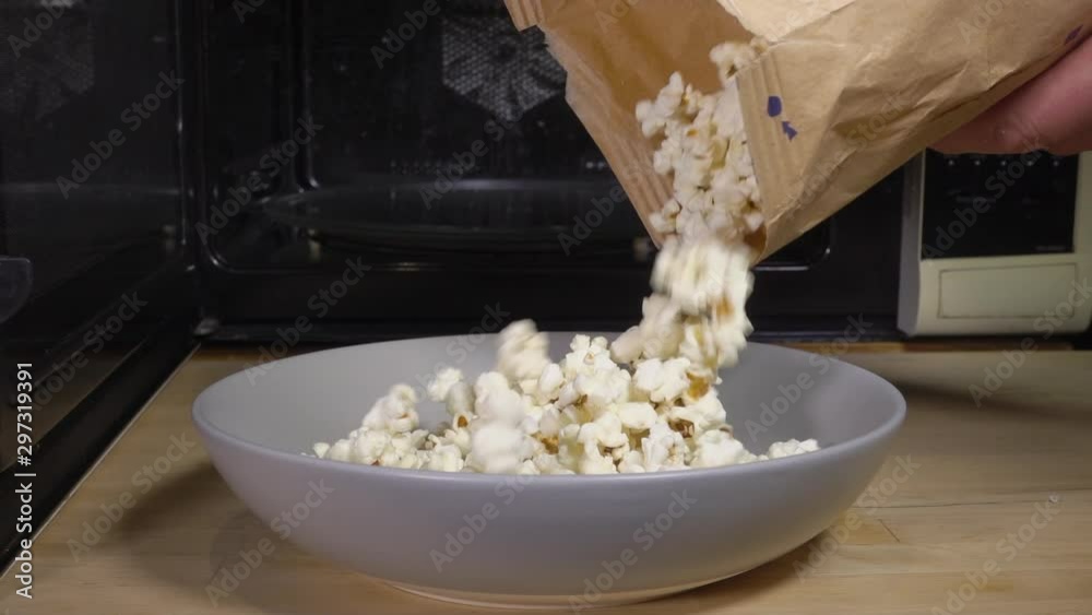 Normal speed, slow motion then normal again. Close POV shot of hot popcorn - which has just been made in the microwave in the background - being poured from the packet into a bowl, ready to eat.