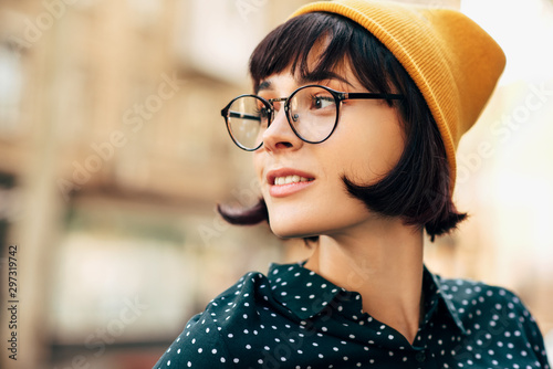 Close-up portrait of beautiful young woman wearing yellow hat and green shirt with white dots, looking aside wainting her boyfriend. Young female university student posing in the city street. photo