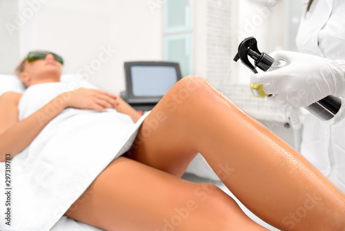 The girl is lying on the couch in the medical glasses in the treatment room. The hands of the cosmetologist make the epilation of the legs
