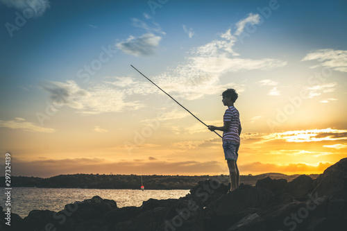 Young fisherman with fishing pole in hand looking the sea on a colorful sunset. Sailor boy with striped t-shirt enjoying a summer evening on the rocks. Lifestyle outdoors fun and carefree concept
