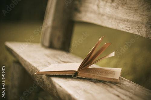 Tablou pe pânză A forgotten open old book lies on a wooden bench cracked with time in the Park in the evening