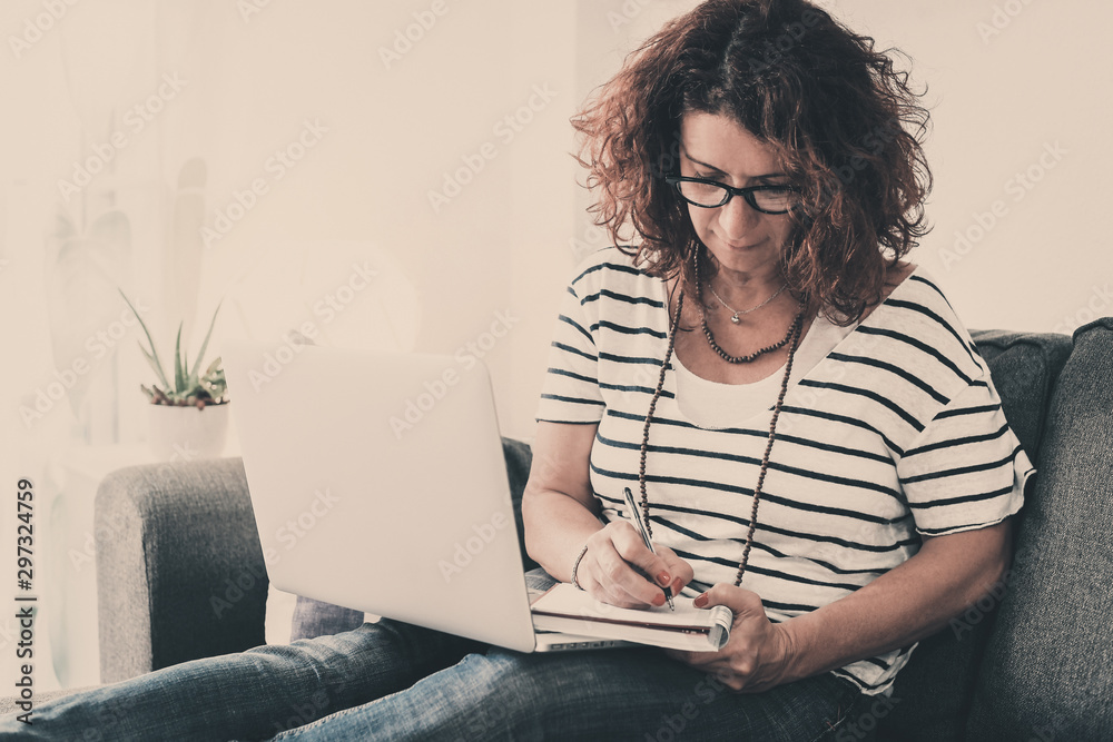 Middle aged woman working at home with laptop sitting on a sofa Mother works part-time in her house with remote access to the office Business girl writing. Home-works new technology lifestyle concept Middle aged woman working at home with laptop sitting on a sofa Mother works part-time in her house with remote access to the office Business girl writing. Home-works new technology lifestyle concept