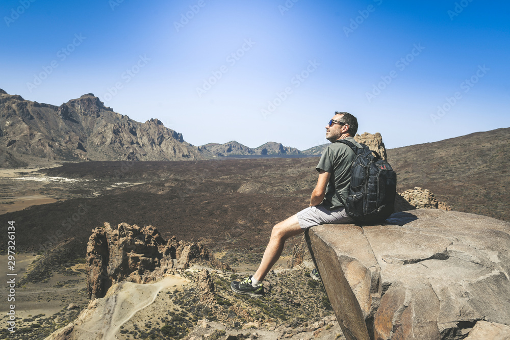 Sporty man with backpack enjoying a beautiful day after climbing alone. Caucasian boy looking away sitting on a mountain cliff overlooking the panorama. Holiday, travel, freedom, outdoor concept.