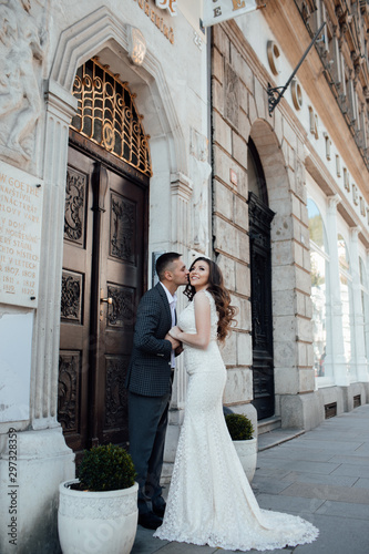 Young romantic couple standing and hugging at city street on a sunny summer day. Handsome guy in suit and girl in white dress with a train. A love story. Beautiful young man and woman pose