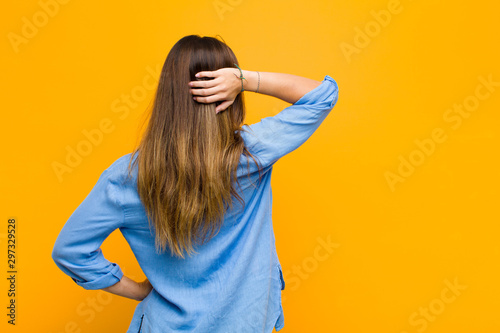young pretty woman thinking or doubting, scratching head, feeling puzzled and confused, back or rear view against orange wall