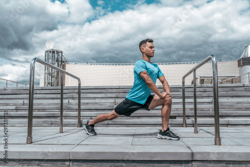 Fototapeta Naklejka Na Ścianę i Meble -  Male athlete doing stretching before training, lunges, jogging before jogging, summer in city, cloud steps background. Active youth lifestyle, fitness workout in nature.
