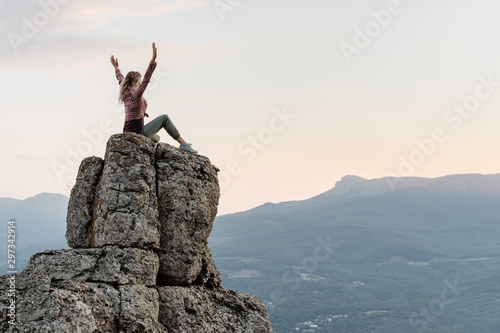 A young woman, glad to have climbed the mountain sits on peak with raised hands and enjoys an amazing view.
